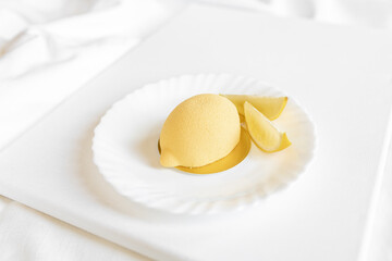 Yellow mousse cake in the form of a lemon lies on a white plate on a light background. Interesting and tasty food. Culinary background