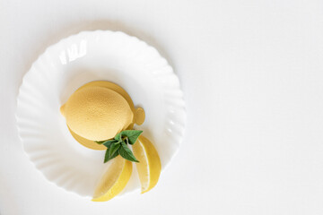Mousse yellow cake in the form of a lemon with a green mint leaf and real lemon slices on a white plate on a light background. Delicious and beautiful pastries. Background for confectionery