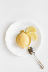 Yellow mousse cake in the form of a lemon lies on a white plate with a spoon on a light background. Interesting and tasty food. Culinary background