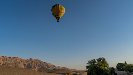 Fototapeta na wymiar A bright yellow balloon is flying in the blue sky. A picturesque mountain range and green trees below. Copy space. Egypt. Luxor