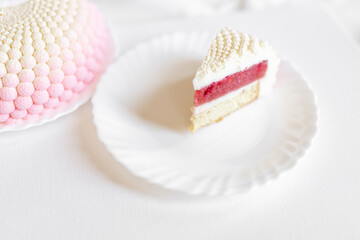 A white-pink piece of cake lies on a white plate on a light background. Delicious pastries