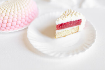A white-pink piece of cake lies on a white plate on a light background. Delicious pastries