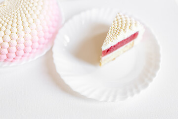 A white-pink piece of cake lies on a white plate on a light background. Delicious pastries