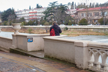 A fisherman is fishing with a fishing rod near the parapet near the river in the city. The fisherman has a red backpack. There are houses on the other side of the river. Selective focus.