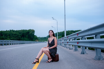 Fototapeta premium A girl with long hair in a black dress is sitting on a suitcase on the side of an empty road and waiting for a car, the concept of hitchhiking