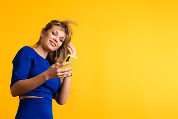 Young woman in blue dress holds modern cell phone, smiles broadly and surprise over yellow background. Positive european girl looking at camera and joy using mobile phone.
