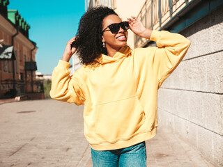 Fototapeta premium Beautiful woman with afro curls hairstyle.Smiling hipster model in yellow hoodie. Sexy carefree female posing on the street background in sunglasses. Cheerful and happy