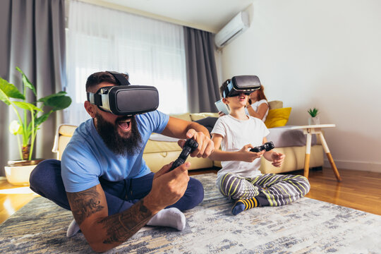 Happy father and son in virtual reality headsets playing with joysticks at home - Powered by Adobe