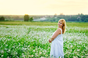 A young plump beautiful woman is resting on a chamomile field at sunset. A plus-size woman in a white sundress in a meadow with daisies .