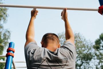 Athletic shaved man pulling himself up on horizontal bar, back view. Male sportsman training on sports ground outdoors during day. Active healthy lifestyle. Selective focus