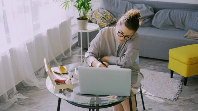 Concentrated woman in glasses watching online lessons on laptop, Lady sitting at table draws in sketchbook in contemporary living room, tracking shot