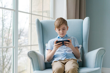 Image of little cheerful boy holding smartphone and playing game while sitting in a chair by the window at home. Look at the phone. Boy with the phone. Cute little boy playing with smartphone