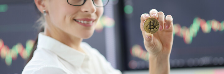 Portrait of young woman with glasses holding bitcoin against background of financial indicators