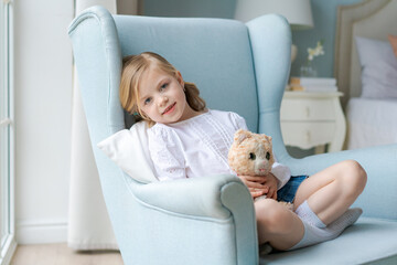 Cute caucasian girl is sitting in blue armchair by window at home in bedroom with a soft toy in her hands and smiling. Happy childhood time. Child spends time with a toy cat