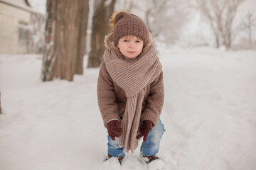 a little handsome boy in brown knitted winter clothes walks along a snow-covered street near the house