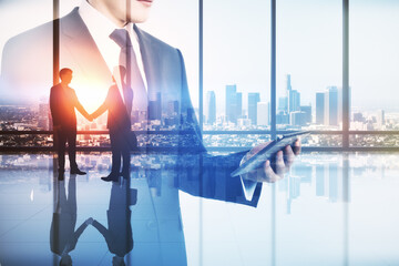 Abstract image of businessman using tablet in modern office interior with panoramic city view, businesspeople shaking hands and mock up place. Teamwork, leadership and technology concept.