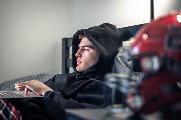 A teenager guy sits in a room on a bed and uses a laptop.