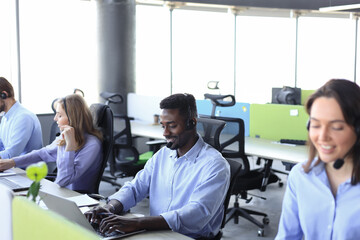 Obraz premium Smiling african male call-center operator with headphones sitting at modern office with collegues on the backgroung, consulting online.