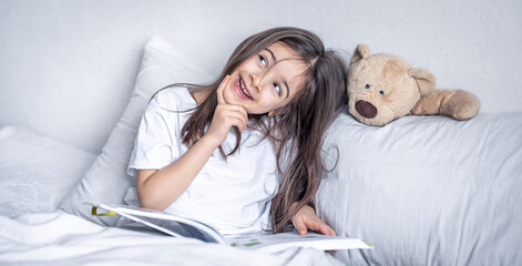 Little girl reads a book with a teddy bear in bed in the morning.
