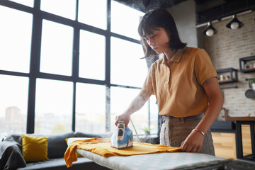 Waist up portrait of young Asian woman ironing clothes at home with steam, household chores concept, copy space