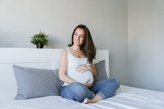 Happy pregnant woman with long brown hair sitting on bed at home