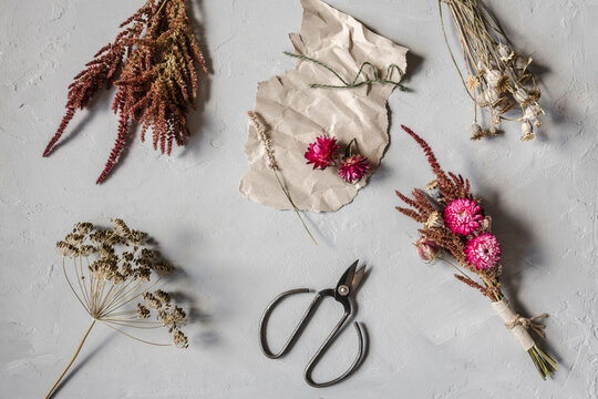 Studio Shot Of Scissors, Piece Of Paper And Various Dried Flowers Flat Laid Against White Background