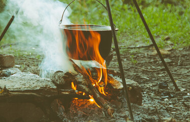 Cooking on an open fire in the field. A camp pot on a campfire close-up on a summer, clear day. Pot on fire with smoke close-up.Cauldron on fire. Cooking dish in a metal pot on an open fire.