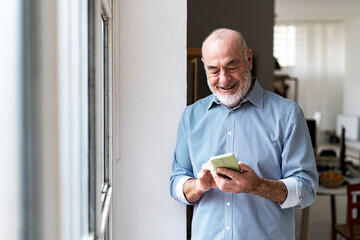 Smiling senior businessman using smart phone by window at home