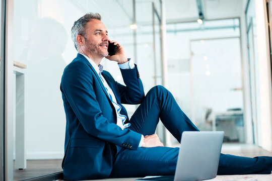 Smiling Businessman Sitting With Laptop Talking On Mobile Phone In Office Corridor