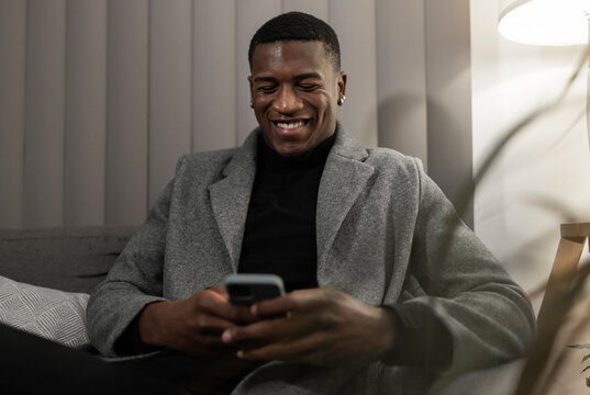 Smiling young man using smart phone sitting on sofa at home