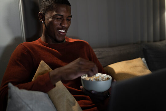 Smiling man watching movie on laptop eating popcorn at home
