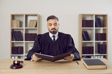 Serious male judge reads legal documents on case sitting at table in his office. Portrait of middle aged man in mantle judge sitting near gavel and court book with folder of documents.