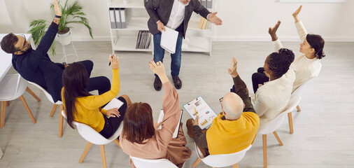Experienced coach engaging people during a business team training. Group of multiracial male and female employees sitting on office chairs, raising hands, willing to participate and answer questions