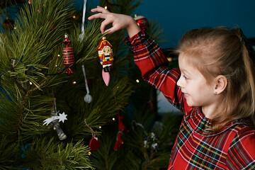 a girl in a red plaid dress hangs a nutcracker toy on the Christmas tree. Christmas Holiday Tree