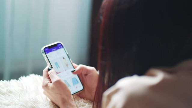 Online shopping concept. Young woman sitting on the bed and makes online shopping on a smartphone. Woman at Home Lying on Couch in Living Room Using Smartphone Buys in Internet Shop.