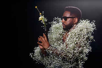 Fashion portrait of a black man in a gypsophila coat holding a pistol shooting a bouquet of flowers.