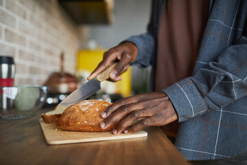Close up of unrecognizable African-American man cutting loaf of fresh homemade bread while cooking in kitchen, copy space