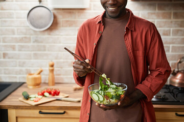Cropped shot of smiling black man mixing salad in glass bowl while enjoying healthy meal in cozy...