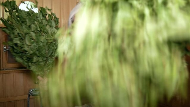 Sauna.
Young man shakes two birch brooms with bitterness and anger. He steams the viewer with brooms in bathhouse.