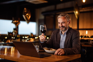Cheerful adult man, joining a conference call on a laptop, while eating at a restaurant.