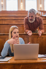 Mature teacher with chalk talking to student near notebooks and devices in university