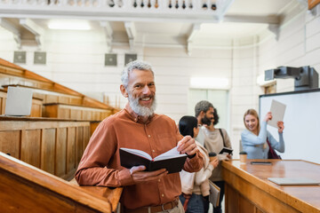 Positive mature teacher holding notebook near blurred students in university auditorium