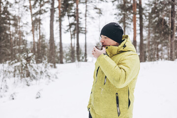 Young man with a cup of tea and thermos in winter snowy forest. White picnic. Drink hot beverage in nature. Local travel. Slow life. Wintertime activity outdoors.