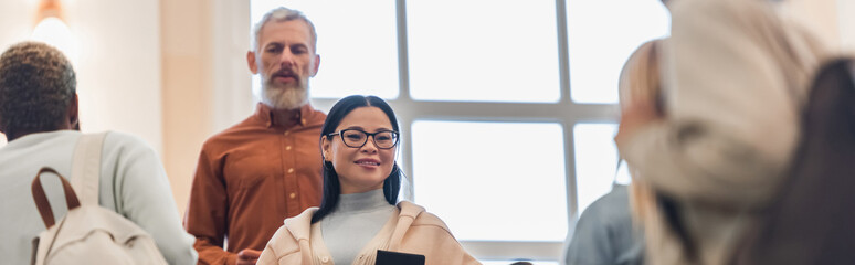 Happy asian student with notebook looking at camera near blurred teacher and interracial friends in...