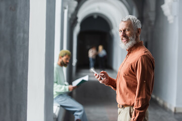 Smiling mature professor using smartphone and looking at camera in university corridor