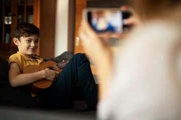 Little boy playing guitar at home. Mother filming her son playing an instrument..