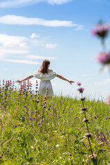 Silhouette of girl in white dress standing in middle of meadow with her arms outstretched against sky. There is blurry plant in foreground. Free life. Selective focus.