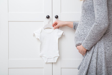 White baby bodysuit hanging on hanger at door of wardrobe at nursery room. Young adult pregnant...