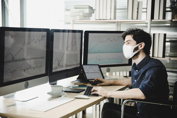 A businessman wears face mask and checks stock market data on monitor computer