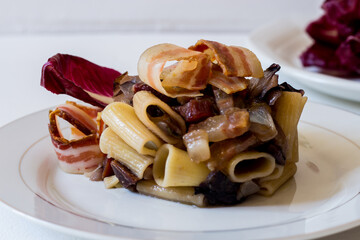 Pasta with bacon and red radicchio, served in a white plate on a white background. Raw radicchio in the background
